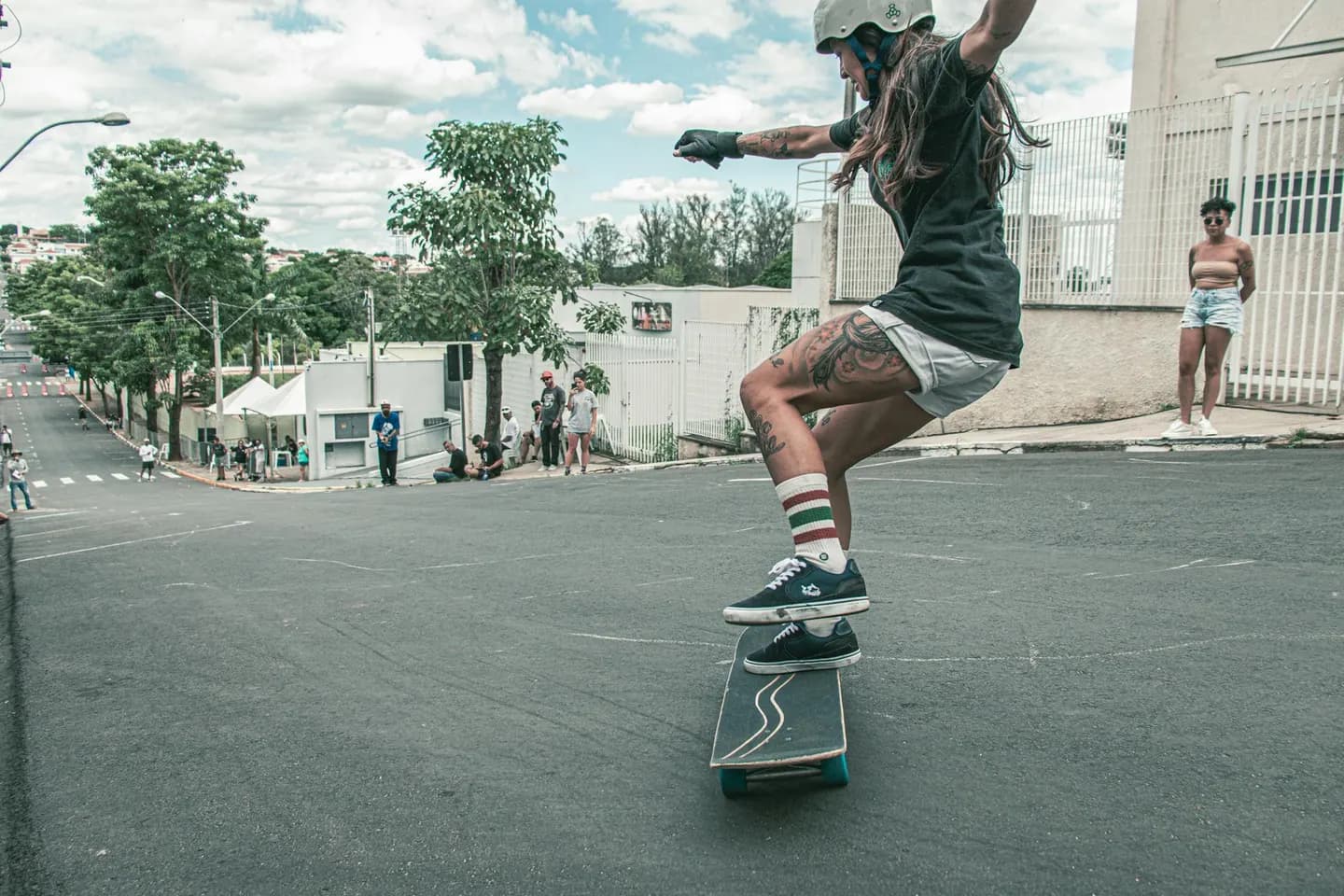 A young woman is skateboarding down a steep, urban street. She is wearing a helmet, black t-shirt, white shorts, and knee-high striped socks. Her arms are outstretched for balance as she maneuvers the skateboard, showcasing her tattoos on her legs. The background features several onlookers, a mix of people standing and sitting by the roadside.